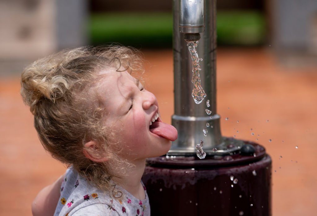 A little girl trying to catch water from a fountain outdoors with her tongue stuck out and her eyes closed.