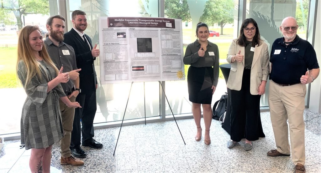 Six people standing on both sides of a capstone project presentation by students and clients. 