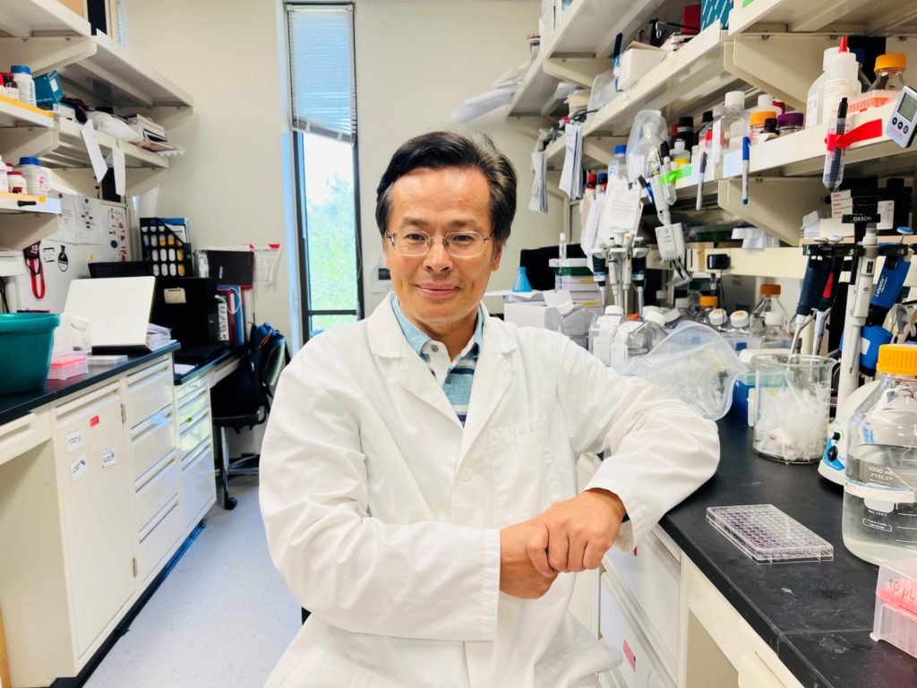 A man in a lab coat, Shaodong Guo, Ph.D., stands in his lab with his equipment behind him.