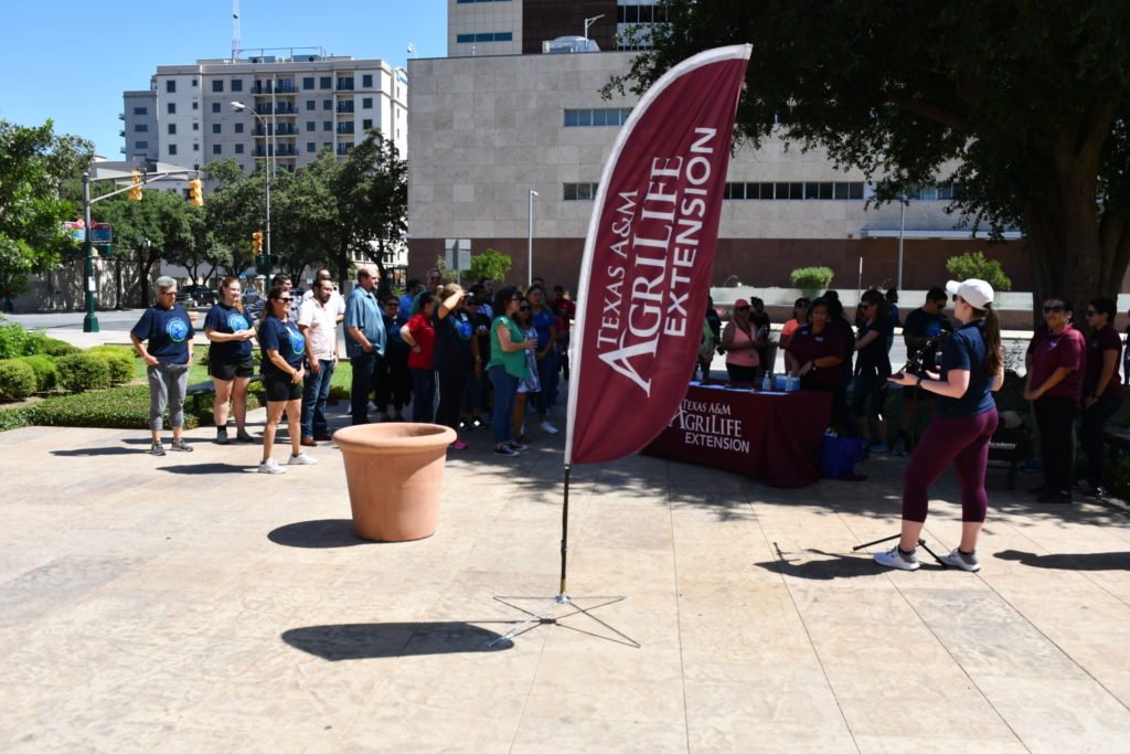 A maroon banner with Texas A&M AgriLife Extension waves on a courtyard area as people are gathered around a table and listening to someone talk about health and wellness