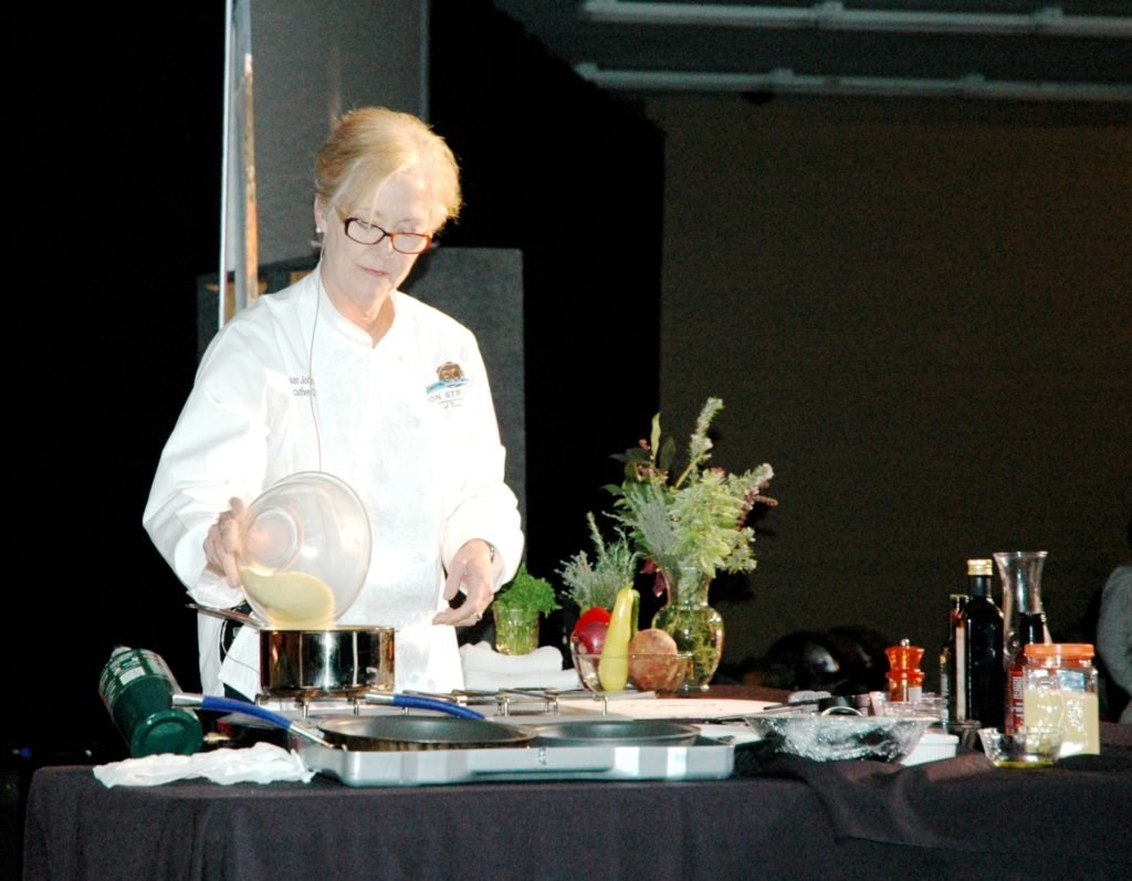 A woman stands at a kitchen counter and pours something out of a bowl into a sauce pan on the stove.