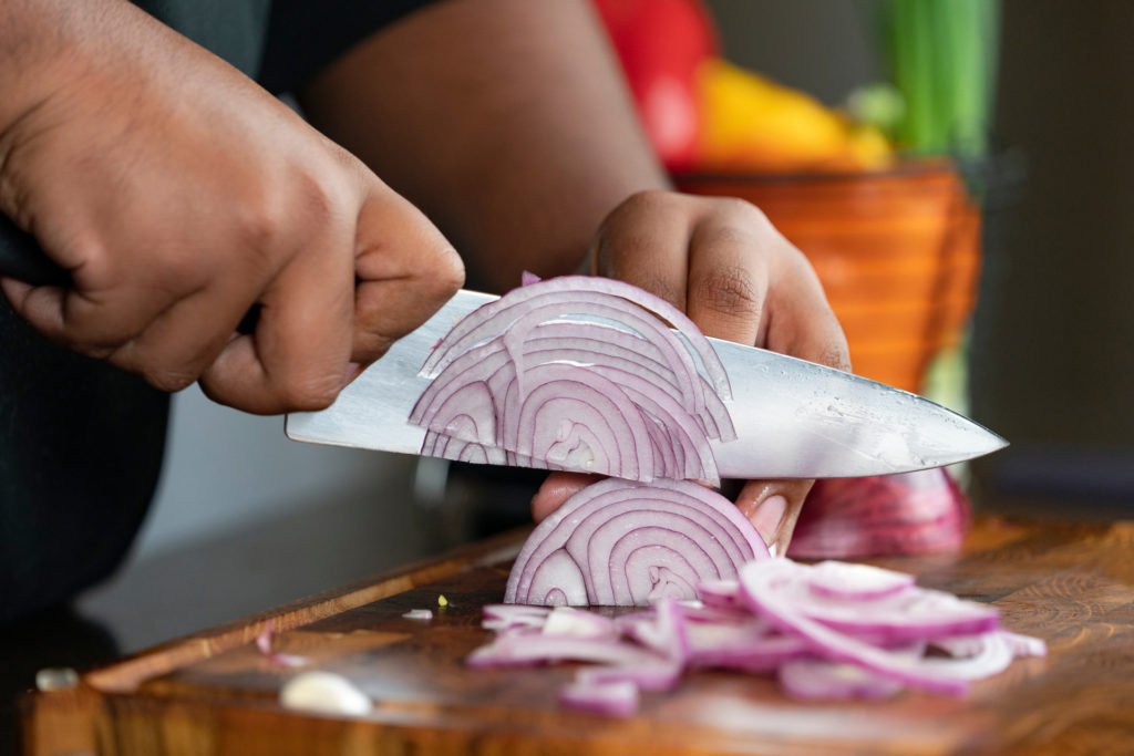 A woman's hands holding a knife cutting a purple onion in thin slices on a cutting board