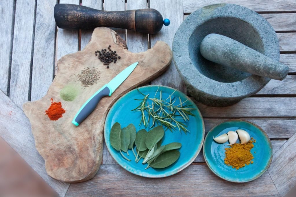 An assortment of spices and herbs on a stone slab amd blue plates. There is also a stone bowl for grinding them up.