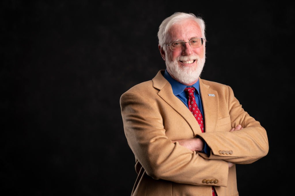 A white-headed man in a red tie, blue shirt and light brown/tan jacket with arms folded -Sandy Pierson 