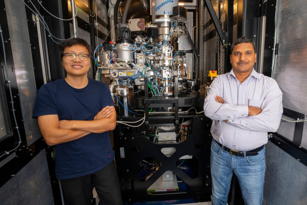 Two men standing cross-armed with complex cryo-EM equipment in the background