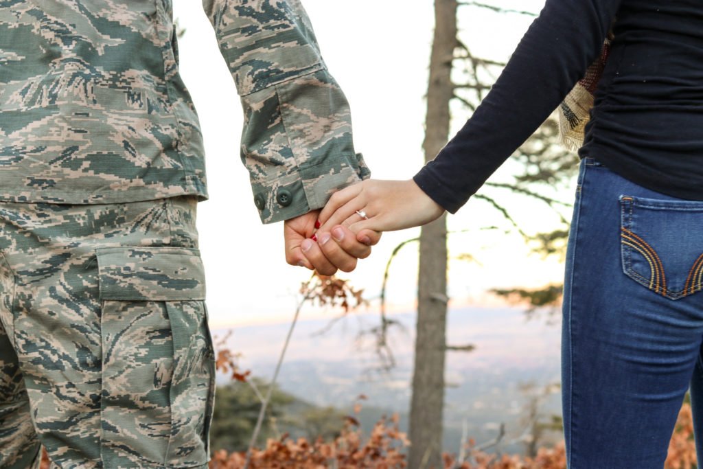 A man in a camouflaged military uniform holds the hand of a woman in jeans and a black long sleeve shirt. We see them from behind from the waist down standing in a wooded area.